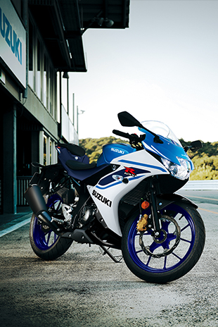 Seitenprofil einer weiß-blauen Suzuki GSX-R125, die auf einem Seitenständer im Fahrerlager einer Rennstrecke steht; im Hintergrund ist das Suzuki-Logo an einem Gebäude zu sehen.