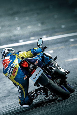 Rückansicht eines Motorradfahrers in blau-gelber Suzuki-Lederkombi, der eine GSX-R125 in Schräglage durch eine Kurve auf der Rennstrecke steuert; Fokus auf das Heck und das Rücklicht.