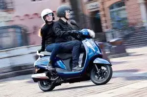 Zwei Personen fahren auf einem blauen Suzuki Address 125 Roller durch eine städtische Umgebung.