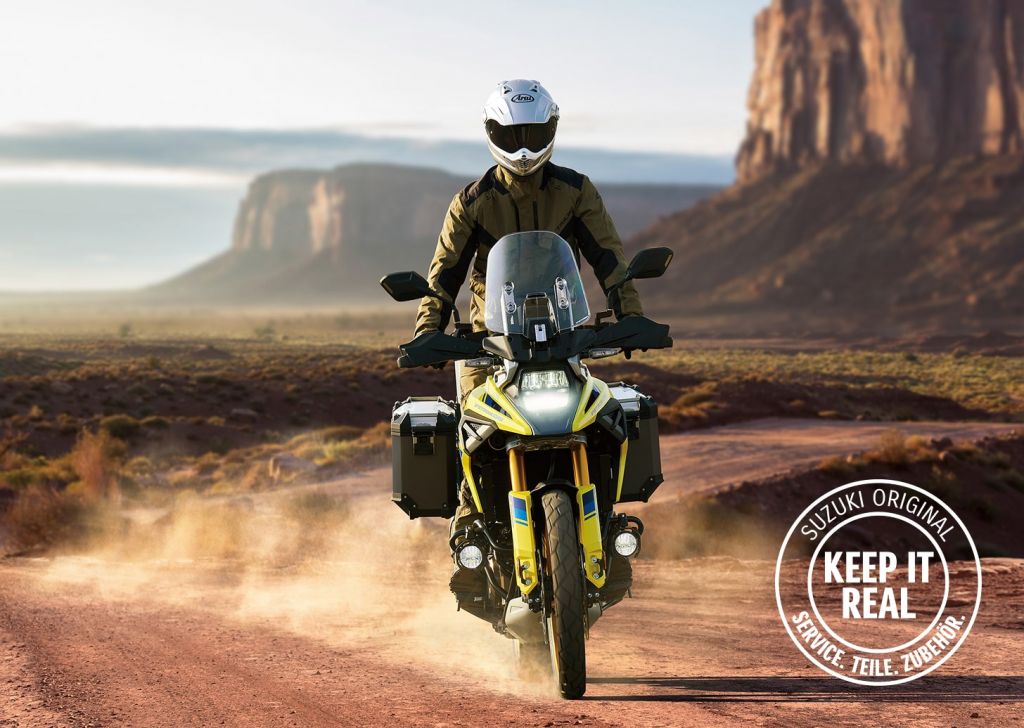 Das Bild zeigt ein Motorrad, das auf einer staubigen Straße in einer landschaftlich reizvollen Umgebung fährt. Das Motorrad ist mit Seitenkoffern ausgestattet und verfügt über zahlreiche weitere Zubehörteile, die einen robusten und abenteuerlichen Charakter unterstreichen. Im rechten unteren Bereich befindet sich ein rundes Logo mit dem Schriftzug „SUZUKI ORIGINAL – KEEP IT REAL – SERVICE. TEILE. ZUBEHÖR.“ Die Szene vermittelt Freiheit, Abenteuer und Authentizität.