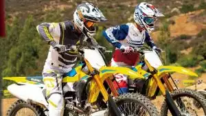 Zwei Suzuki RM-Z250 Motocross-Fahrer in voller Rennmontur starten dynamisch auf staubiger Strecke.