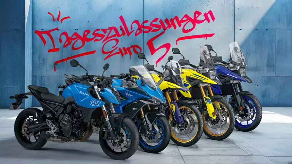 5 verschiedene Suzuki Motorradmodelle auf grauem Betonboden, leicht versetzt nebeneinander angeordnet. Eine GSX-8S und eine GSX-S1000GT in hellblauer Farbe, eine V-Strom 800DE und eine V-Strom 1050DE in gelber Farbe und eine V-Strom 1050 in dunkelblauer Farbe. Im Hintergrund eine hellblaue Wand mit Schriftzug im Graffiti-Look: "Tageszulassungen/Euro 5".